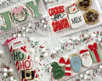 May include: Assortment of Christmas-themed sugar cookies in clear packaging. Cookies feature festive designs like "JOY", "Cookies for Santa", "Milk", and "Ho Ho Ho".