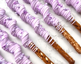 Puede incluir: Primer plano de un grupo de palitos de pretzel cubiertos de chocolate morado y decorados con chispas moradas. Los pretzels están dispuestos en una fila sobre una superficie de mármol blanco.