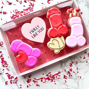 May include: A set of three Valentine's Day cookies decorated with pink, red, and white icing. The cookies are shaped like hearts and have the text "I WILLY LIKE YOU" on one of the cookies.