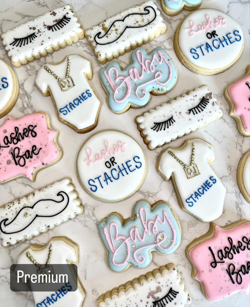 May include: A collection of decorated sugar cookies with various designs, including eyelashes, mustaches, baby onesies, and the words "Lashes or Staches" and "Baby".