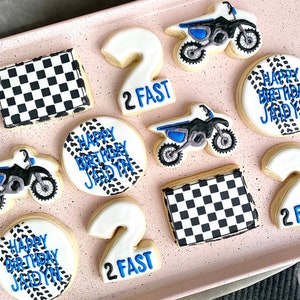 Pu&ograve; includere: Un set di dieci biscotti di zucchero decorati con un tema di motocross. I biscotti sono decorati con glassa blu e bianca e includono bandiere a scacchi, il numero due e le parole "Buon compleanno Jadyn" e "2 Fast".