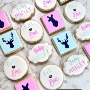 May include: A collection of decorated sugar cookies with various designs, including a deer silhouette, the words "Baby Hadley", and "Buck or Doe". The cookies are decorated with pink, blue, and white icing.
