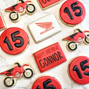 Pu&ograve; includere: Un set di biscotti di zucchero decorati in rosso e bianco con un tema motocross. I biscotti presentano il numero 15, le parole "Happy Birthday Connor" e un logo Honda.