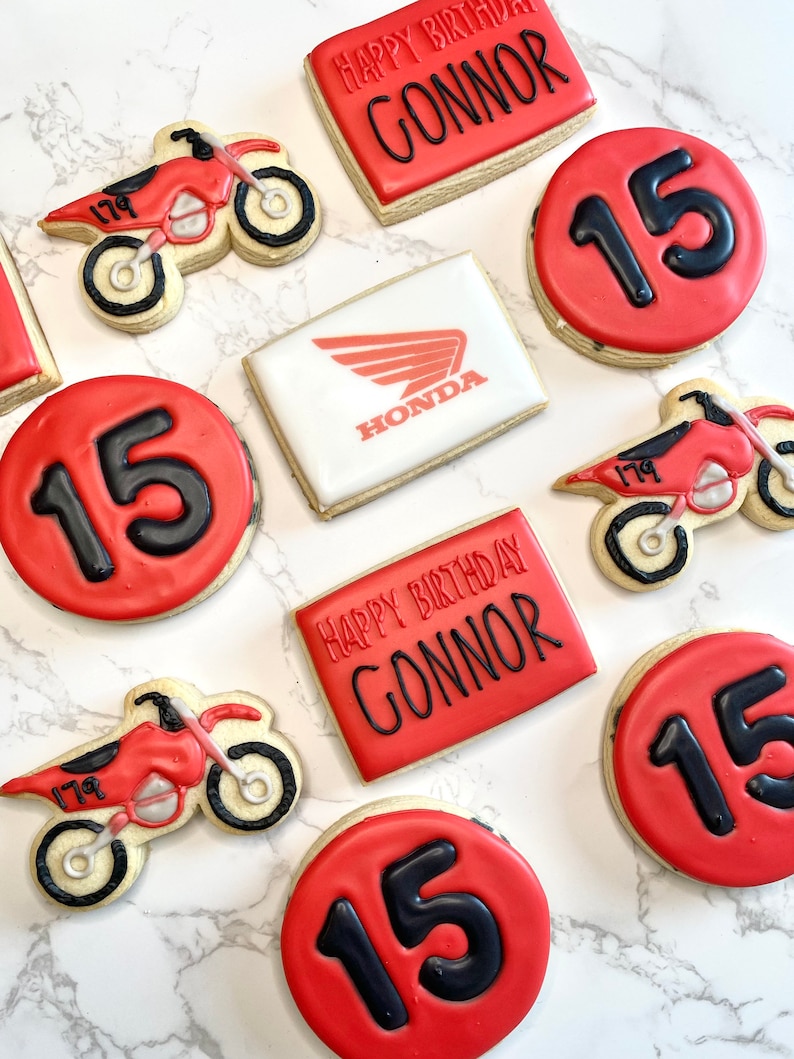 Pu&ograve; includere: Un set di biscotti di zucchero decorati in rosso e bianco con un tema motocross. I biscotti presentano il numero 15, le parole "Happy Birthday Connor" e un logo Honda.