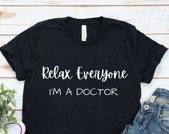 May include: A black t-shirt with the text "Relax Everyone I'm A Doctor" in white lettering. The shirt is laid flat on a white wood surface with a pair of white trainers and blue jeans.
