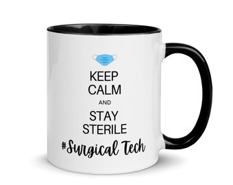 Puede incluir: Taza de café de cerámica blanca con borde y asa negros. La taza tiene un gráfico de mascarilla facial azul y blanca y el texto "Keep Calm and Stay Sterile #Surgical Tech".