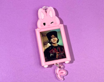 May include: A pink acrylic keychain featuring a photo within a rectangular frame. The frame is topped with a winking bunny head. A pink swirl charm hangs below the frame. The background is a solid purple color.