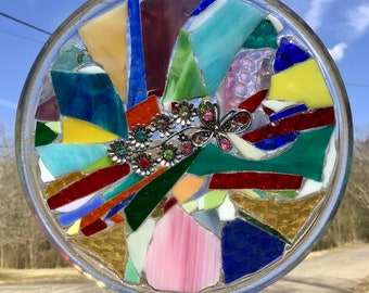 Könnte beinhalten: Ein runder, transparenter Glassonnenfänger mit einem Mosaikdesign aus bunten Glassplittern. Das Mosaik zeigt einen Schmetterling mit Strasssteinen und grünen und rosa Blumen.
