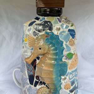 May include: A ceramic bottle with a mosaic design featuring a seahorse, seashells, and palm trees. The bottle has a wooden lid and a handle.
