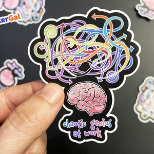 May include: A die-cut sticker featuring a pink human brain illustration at the bottom with a tangle of colorful arrows and lines above it on a black background. The text "chaotic genius at work" is printed in a playful, multi-colored font below the brain.