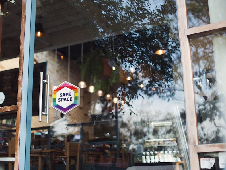 May include: A glass door with a rainbow-colored sticker that says "SAFE SPACE" on it. The sticker is a hexagon shape.