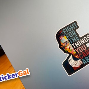 May include: A die-cut sticker featuring a colorful, stylized portrait of Ruth Bader Ginsburg wearing her signature lace jabot. The text "FIGHT FOR THE THINGS YOU CARE ABOUT" is printed in bold, block letters next to the portrait on a dark background.