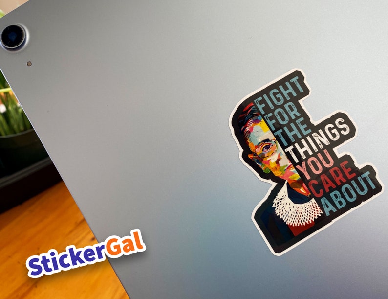 May include: A die-cut sticker featuring a colorful, stylized portrait of Ruth Bader Ginsburg wearing her signature lace jabot. The text "FIGHT FOR THE THINGS YOU CARE ABOUT" is printed in bold, block letters next to the portrait on a dark background.
