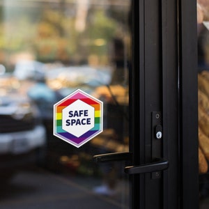 May include: A hexagonal sticker with a rainbow border and the words "SAFE SPACE" in white text on a white background. The sticker is on a glass door.