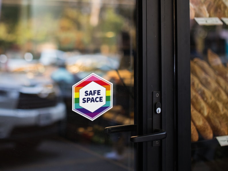 May include: A hexagonal sticker with a rainbow border and the words "SAFE SPACE" in white text on a white background. The sticker is on a glass door.