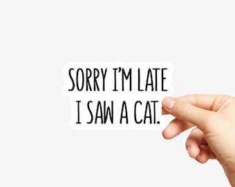 May include: A white die-cut sticker featuring the black text SORRY I'M LATE I SAW A CAT. The sticker has a clean, simple font and a white border, presenting a humorous message about being distracted by a feline in a clear, legible format.