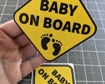 May include: A diamond-shaped yellow sticker with a black border featuring the text BABY ON BOARD in bold black capital letters above a graphic of two small black baby footprints. A second identical square sticker is positioned below the first one.