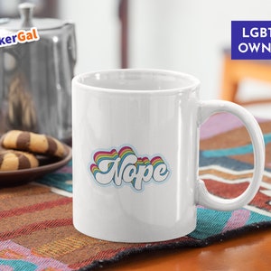 May include: White ceramic coffee mug with a rainbow-colored "Nope" graphic. The mug has a black handle and is sitting on a colorful rug. The text "LGBTQ OWNED" is visible in the background.