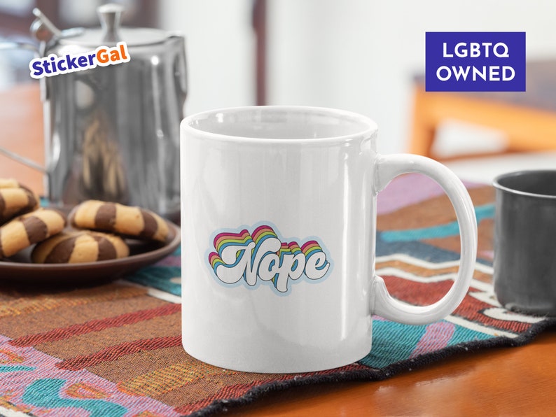 May include: White ceramic coffee mug with a rainbow-colored "Nope" graphic. The mug has a black handle and is sitting on a colorful rug. The text "LGBTQ OWNED" is visible in the background.