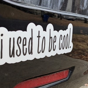 May include: A white vinyl decal with a black scalloped border and the black text "i used to be cool." in a handwritten font. The sticker is applied to a smooth, dark, reflective surface, featuring a clean, die-cut shape around the lettering.