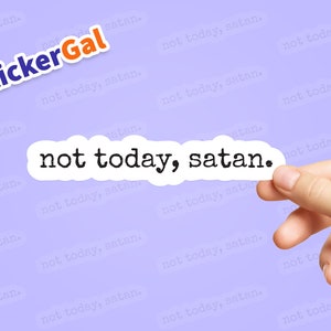 May include: A white sticker with the text "not today, satan." printed in black typewriter font. The sticker is being held by a hand against a light purple background with the text "not today, satan." repeated in a smaller font.