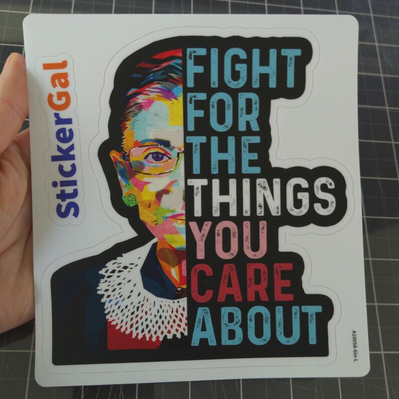 May include: A rectangular sticker featuring a colorful, pop-art style portrait of Ruth Bader Ginsburg on the left side, wearing a white lace jabot. The right side displays the text FIGHT FOR THE THINGS YOU CARE ABOUT in bold, distressed lettering.