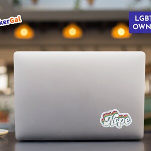 May include: A silver laptop computer with a rainbow sticker that says "Nope" on the back. The laptop is on a brown wooden table.