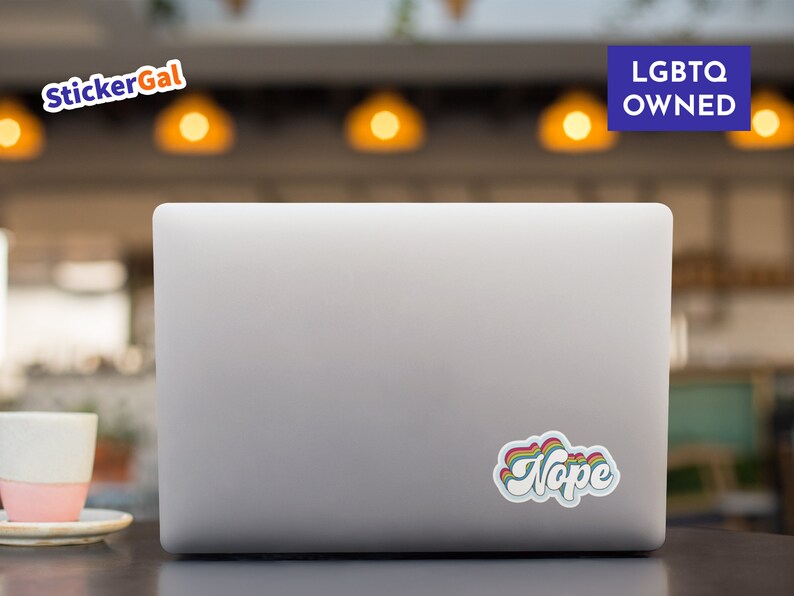 May include: A silver laptop computer with a rainbow sticker that says "Nope" on the back. The laptop is on a brown wooden table.