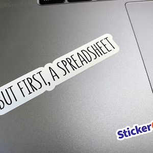 May include: A white rectangular sticker with rounded corners featuring the black text "BUT FIRST, A SPREADSHEET" in a clean sans-serif font. The sticker is positioned diagonally on a metallic gray surface, with a small "StickerGal" logo in the corner.