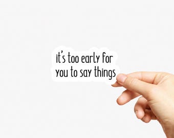 May include: A white die-cut sticker featuring the black text 'it's too early for you to say things' in a simple, rounded sans-serif font. The sticker has a clean white border around the lettering, creating a distinct shape against the plain background.