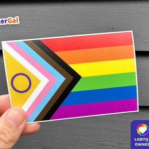 May include: A sticker featuring the Progress Pride flag with a purple circle and a yellow background. The flag has the traditional rainbow colors, plus black and brown stripes, and a light blue, pink, and white chevron.