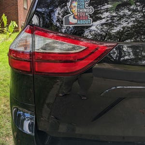May include: A rectangular vinyl sticker applied to the rear window of a black vehicle. The sticker features a colorful portrait of Ruth Bader Ginsburg and the white text "FIGHT FOR THE THINGS YOU CARE ABOUT" with a white border outline.