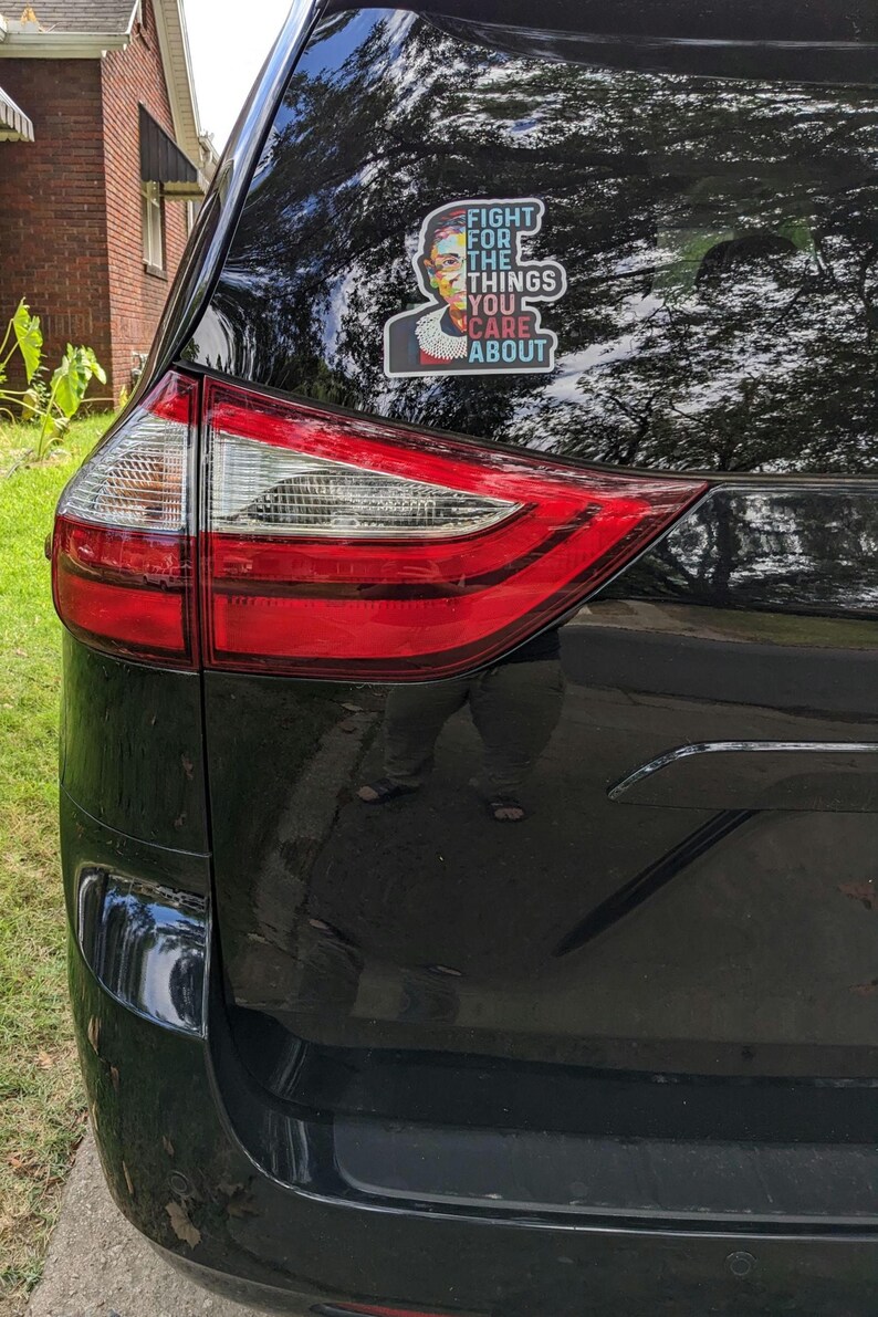 May include: A rectangular vinyl sticker applied to the rear window of a black vehicle. The sticker features a colorful portrait of Ruth Bader Ginsburg and the white text "FIGHT FOR THE THINGS YOU CARE ABOUT" with a white border outline.