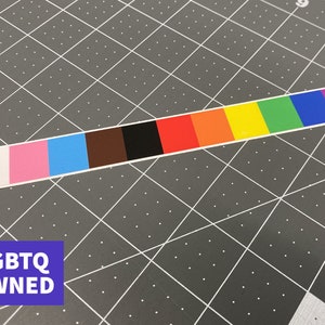 May include: A rectangular sticker with a color gradient representing the LGBTQ+ Pride flag. The sticker features a white, pink, light blue, brown, black, red, orange, yellow, green, dark blue, and purple color scheme. The text "LGBTQ OWNED" is in a blue rectangle.