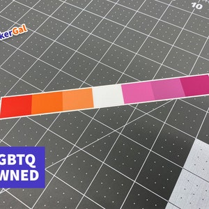 May include: A rectangular sticker featuring a horizontal gradient of seven distinct color bands, transitioning from dark red, orange, light orange, white, light pink, medium pink, to dark pink. The sticker is positioned on a dark gray cutting mat.