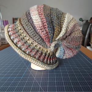 Op de afbeelding: Een gehaakte muts met een veelkleurig swirl-patroon in tinten bruin, beige, roze en blauw. De muts is gemaakt met verschillende steken en texturen, wat een uniek en opvallend ontwerp creëert.