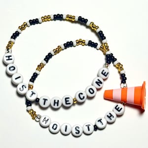 May include: A beaded necklace with the words "HOIST THE CONE" spelled out in white letter beads. The necklace features black, gold, and white beads, and an orange and pink traffic cone charm.