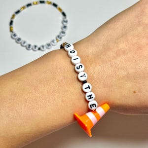 May include: A white beaded bracelet with black letters spelling "HOIST THE" and an orange traffic cone charm. Another bracelet with white and black beads is in the background.