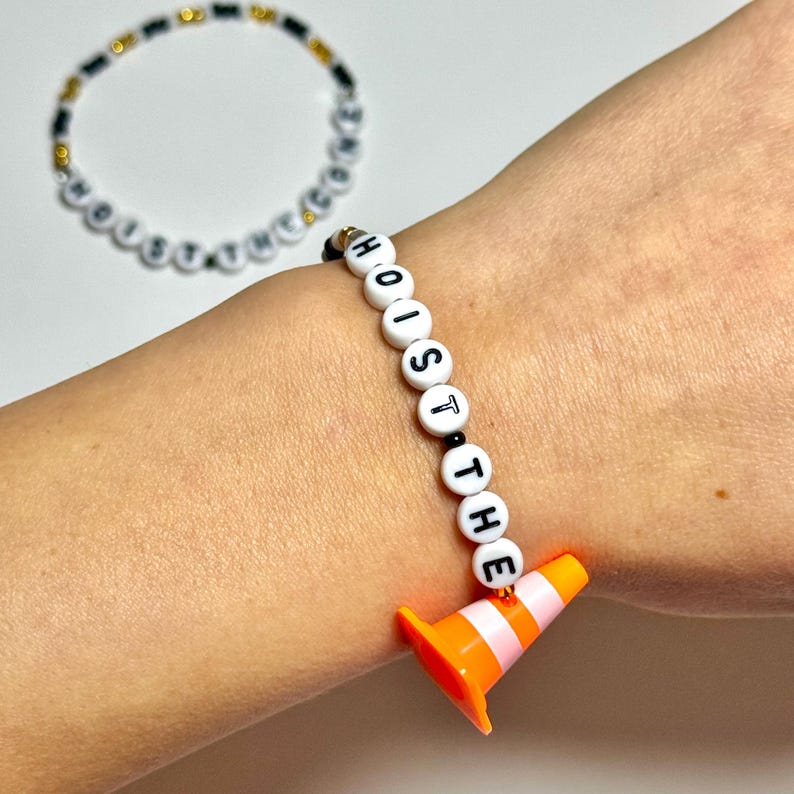 May include: A white beaded bracelet with black letters spelling "HOIST THE" and an orange traffic cone charm. Another bracelet with white and black beads is in the background.