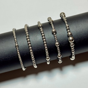 May include: Five silver-toned beaded bracelets arranged in a row. Each bracelet features a unique pattern of metallic spheres, ranging from uniform small beads to alternating sizes of larger and smaller polished round beads on a flexible cord.