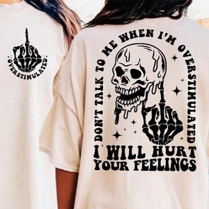 May include: A white t-shirt with a black skull and skeleton hand graphic on the back. The graphic says "Don't talk to me when I'm overstimulated. I will hurt your feelings."