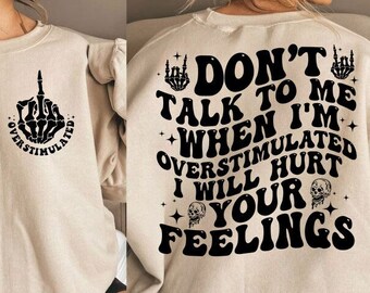 May include: A beige sweatshirt with a black graphic print that says "Don't talk to me when I'm overstimulated I will hurt your feelings". The graphic also includes a skeleton hand giving the middle finger and a skull.