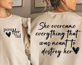 May include: Beige sweatshirt with black text that reads "She overcame everything that was meant to destroy her" with a black heart on the right side of the text. The left side of the sweatshirt has black text that reads "Powerful" with a black heart.