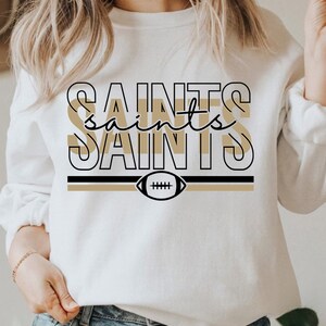 May include: White sweatshirt with a black and gold graphic design featuring the word "Saints" repeated three times with a football in the center.