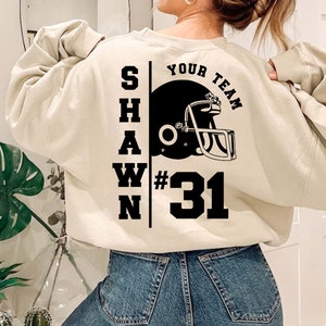 K&ouml;nnte beinhalten: Ein cremefarbener Pullover mit schwarzem Text, der "SHAWN YOUR TEAM #31" lautet, und einer schwarz-wei&szlig;en Illustration eines Footballhelms.