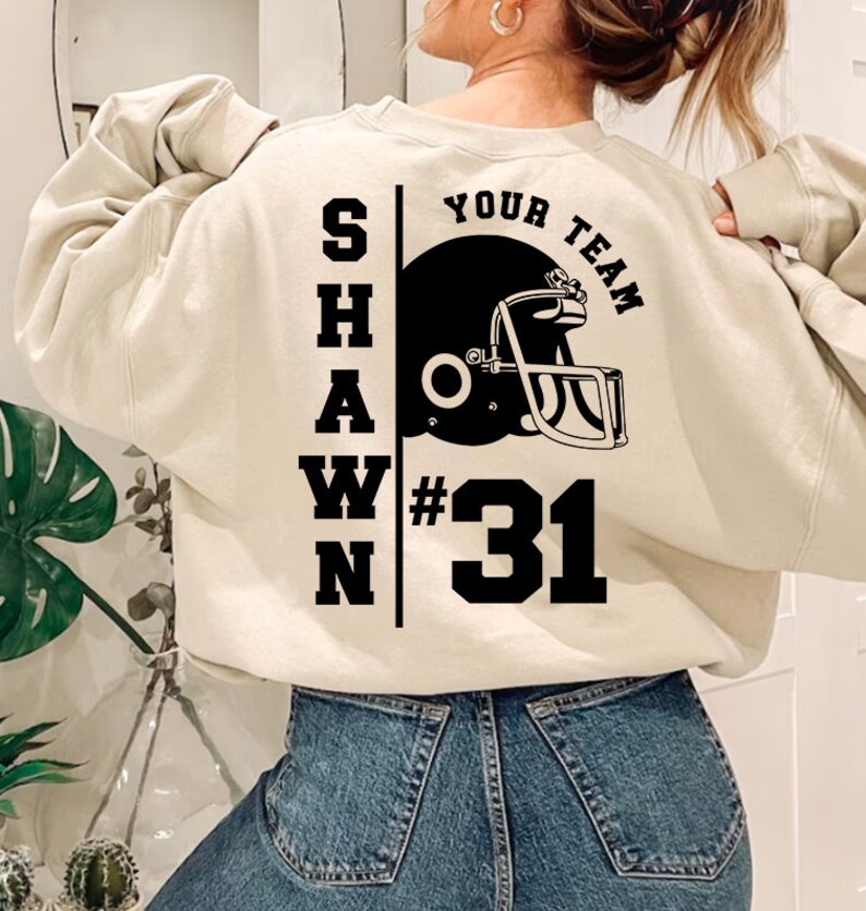 K&ouml;nnte beinhalten: Ein cremefarbener Pullover mit schwarzem Text, der "SHAWN YOUR TEAM #31" lautet, und einer schwarz-wei&szlig;en Illustration eines Footballhelms.