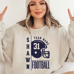May include: A white sweatshirt with a navy blue graphic of a football helmet with the text "TEAM NAME" above the helmet and "SHAWN FOOTBALL" below the helmet. The number "31" is on the helmet.