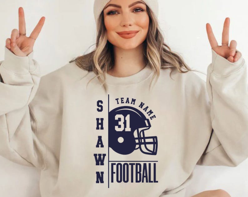May include: A white sweatshirt with a navy blue graphic of a football helmet with the text "TEAM NAME" above the helmet and "SHAWN FOOTBALL" below the helmet. The number "31" is on the helmet.