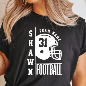 May include: Black t-shirt with white text that reads "SHAWN" and "TEAM NAME" with a football helmet graphic and the number "31". The text "FOOTBALL" is also printed in white.
