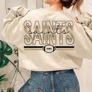 May include: A beige sweatshirt with a black and brown graphic design that says "Saints" with a football graphic underneath.